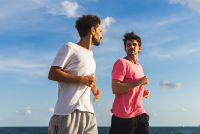 Hispanic male athletes looking at each other and jogging on embankment against cloudy blue sky and sea during cardio workout on sunny day