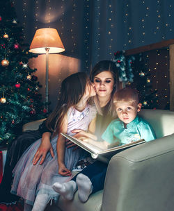 Portrait of boy with sister and mother sitting on chair with book against christmas tree at home