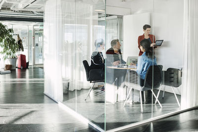 Businesswoman showing digital tablet to colleagues seen through glass