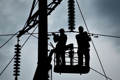 Silhouette workers on cherry picker by electricity pylon against sky