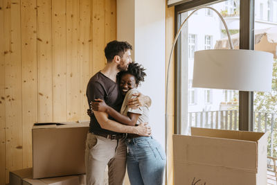 Happy couple embracing each other while standing near window at new home