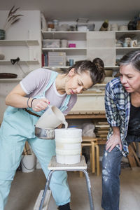 Mature female potter bending while looking at young employee pouring clay from pitcher in vase at workshop