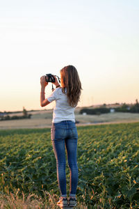 Rear view of young woman photographing through camera while standing on grassy field during sunset