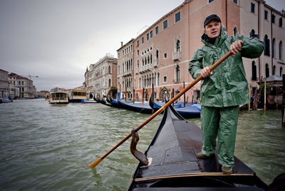 Low angle view of man on gondola in canal against buildings during rainy season