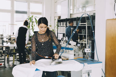 Female entrepreneur ironing white fabric while colleagues working in background at creative office