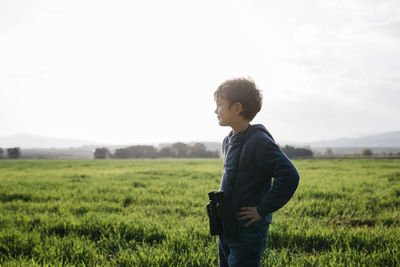 Cute boy with binoculars looking away while standing in field during sunny day