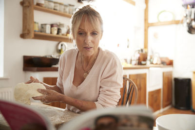 Senior woman kneading while reading recipe book sitting in kitchen