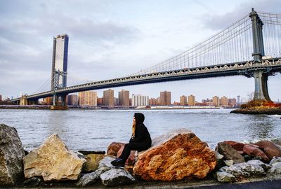 Young woman looking away while sitting on rock by river in city