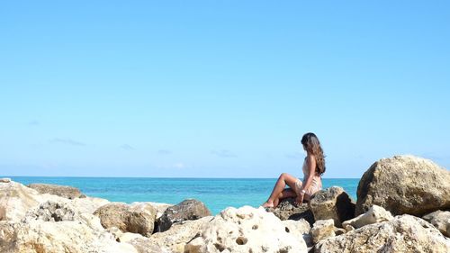 Woman looking at view of sea while sitting on rocks against sky