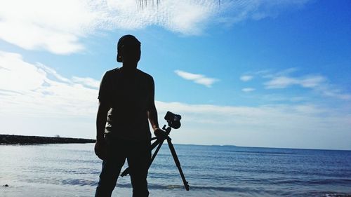 Man with tripod camera standing on sea shore at beach against sky