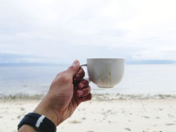 Close-up of hand holding drink at beach against sky