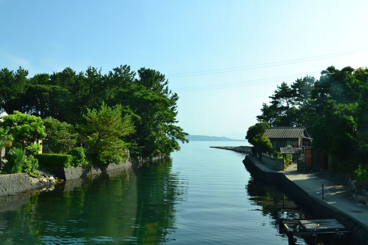 water, tree, clear sky, tranquil scene, scenics, river, architecture, tranquility, building exterior, sea, calm, blue, waterfront, growth, day, nature, beauty in nature, outdoors, riverbank, canal, no people, tourism, in front of, town, lakeside, remote