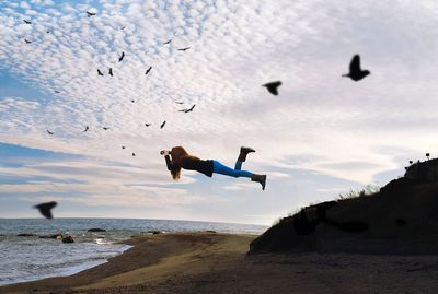 Full length of young woman levitating amidst birds at beach against sky