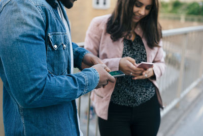 Midsection of man standing by woman while using smart phones on footbridge in city