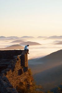 Man sitting on cliff with mountains in background against sky