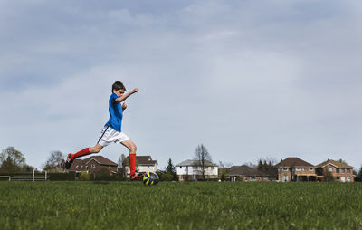 Side view of boy kicking soccer ball while playing on grassy field against sky