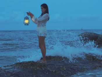 Side view of young woman with illuminated oil lamp standing at beach against sky