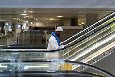 African american passenger man with suitcase stands on escalator, holds handrail in airport terminal