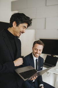 Happy businessman holding laptop by male colleague in office