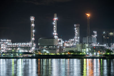 Illuminated factory by river against sky at night