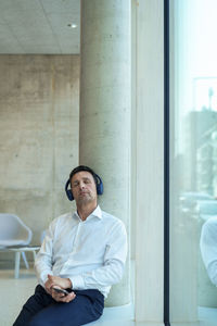 Businessman with headphones sitting by column near window at office