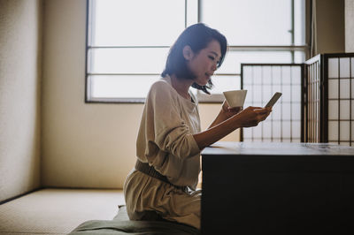 Woman drinking coffee while using smart phone sitting by table at home