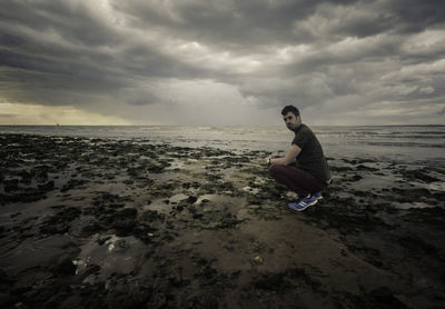 Man crouching on shore at beach against sky
