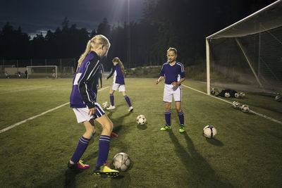 Girls practicing with soccer balls by goal post on field at night