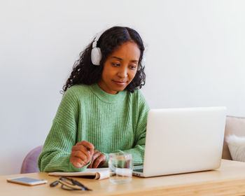 Young african-american female working on laptop in headphones while taking notes in notebook