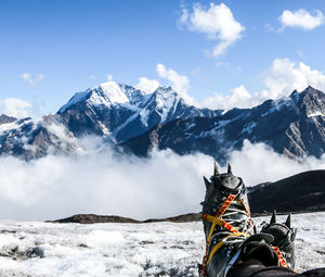 Low section of person wearing snow boot on snow covered field by mountains against sky