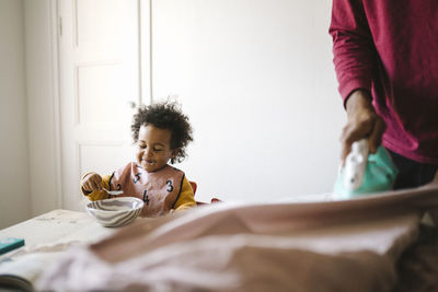 Girl eating from bowl while father ironing clothes