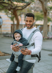 Content african american man embracing son in baby carrier while surfing mobile phone on city street