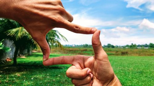 Cropped hands of people making finger frame over field against sky