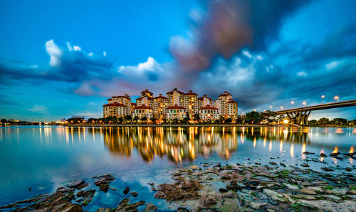 Illuminated buildings by river against cloudy sky