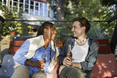 Smiling multiracial male and female holding drink bottles and talking with each other while sitting on bench at party