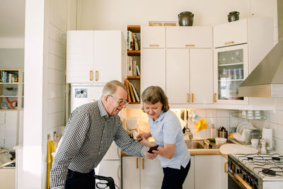 Retired senior man showing smart phone to female volunteer washing dishes in kitchen at nursing home