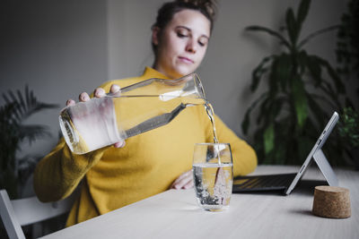 Woman pouring water in glass while sitting with digital tablet at home office