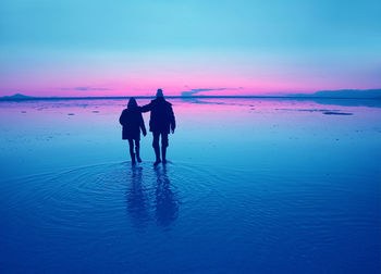 Rear view of couple walking on shore against sky during sunset