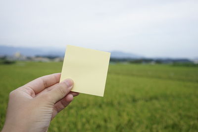 Close-up of hand holding paper on field against sky