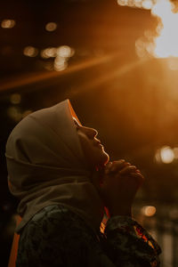 Side view of young woman praying while standing outdoors during sunset