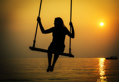 Silhouette girl on swing at beach against sky during sunset