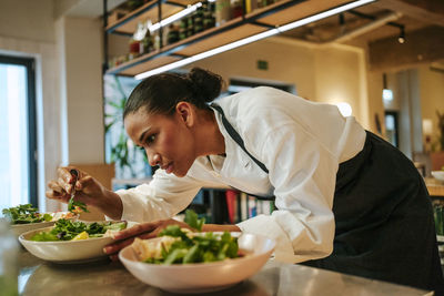 Focused female chef arranging cilantro on salad bowl using tweezers in commercial kitchen