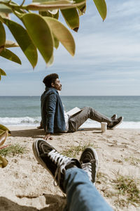 Relaxed man with book sitting on sand at beach during sunny day