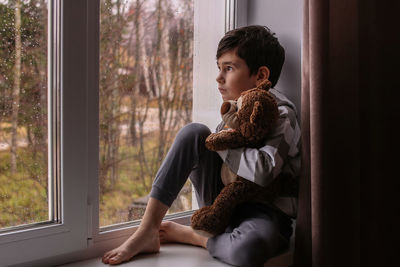 The boy dreamily looks out the window, hugging a teddy bear