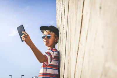 Side view of boy taking selfie from phone while standing by wall