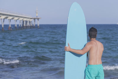 Rear view of shirtless man standing with surfboard at beach