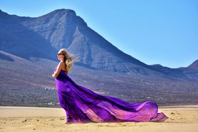 Woman holding purple fabric walking on sand against sky