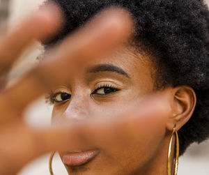 Young african american female with curly hair and hoop earrings outstretching hand and looking at camera while standing against urban building