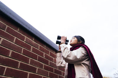 Woman holding binoculars standing near brick wall