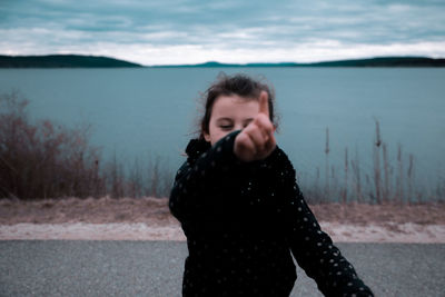 Girl pointing while standing on road by lake against cloudy sky during sunset